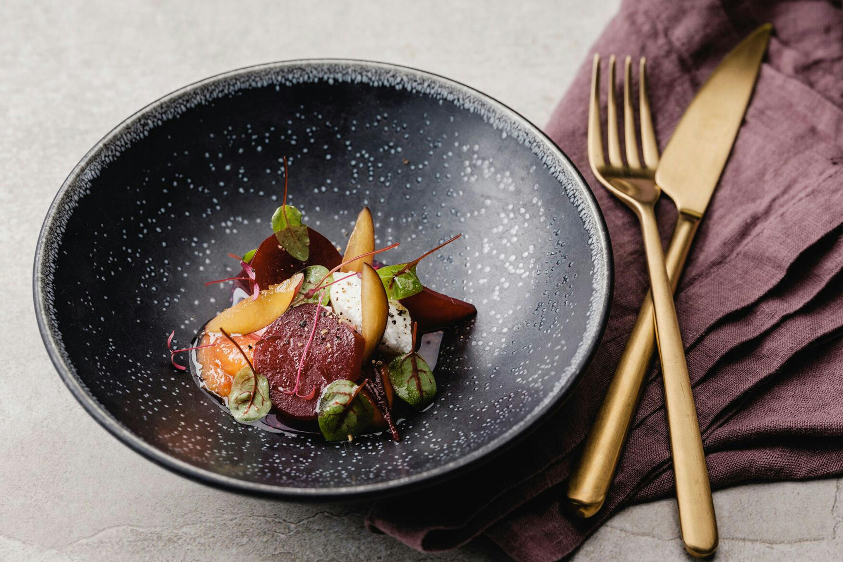 Lukewarm beets with caramelized plums and fresh goat's cheese in a black deep plate with gold cutlery