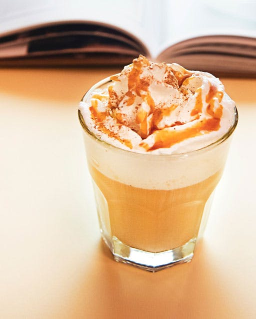 a cup of milky coffee with whipped milk in a glass besides an open book