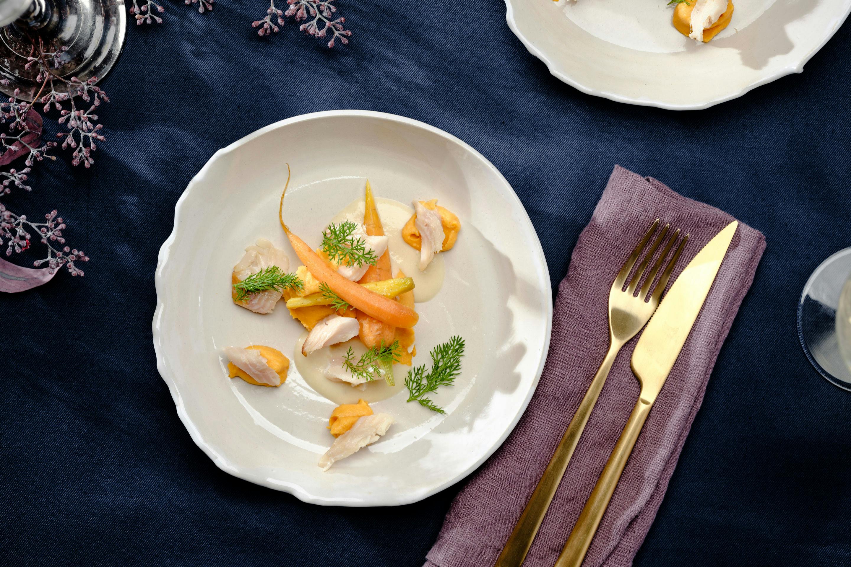 Two kinds of field carrots with carrot puree and smoked trout fillet pieces on a cream-colored plate, arranged with golden cutlery on a dark blue tablecloth