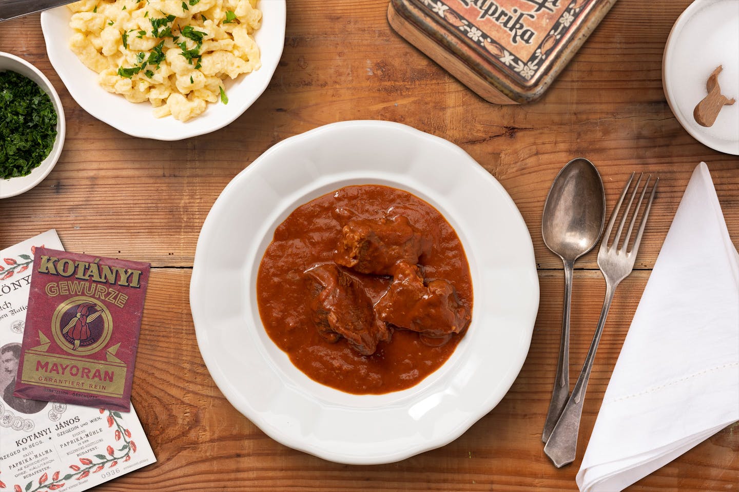 A plate full of goulash and a small bowl of spaetzle arranged on a dark wooden table.