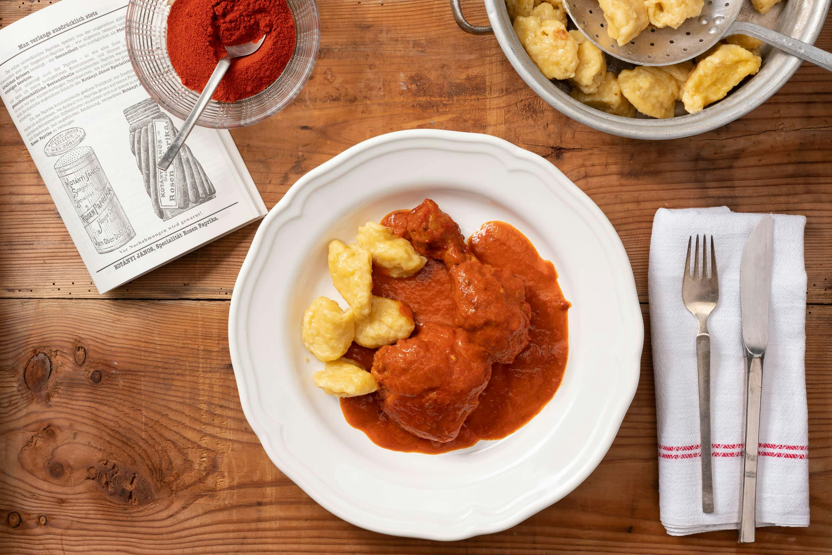 A deep plate with paprika chicken and dumplings, beside which is a bowl full of ground paprika.