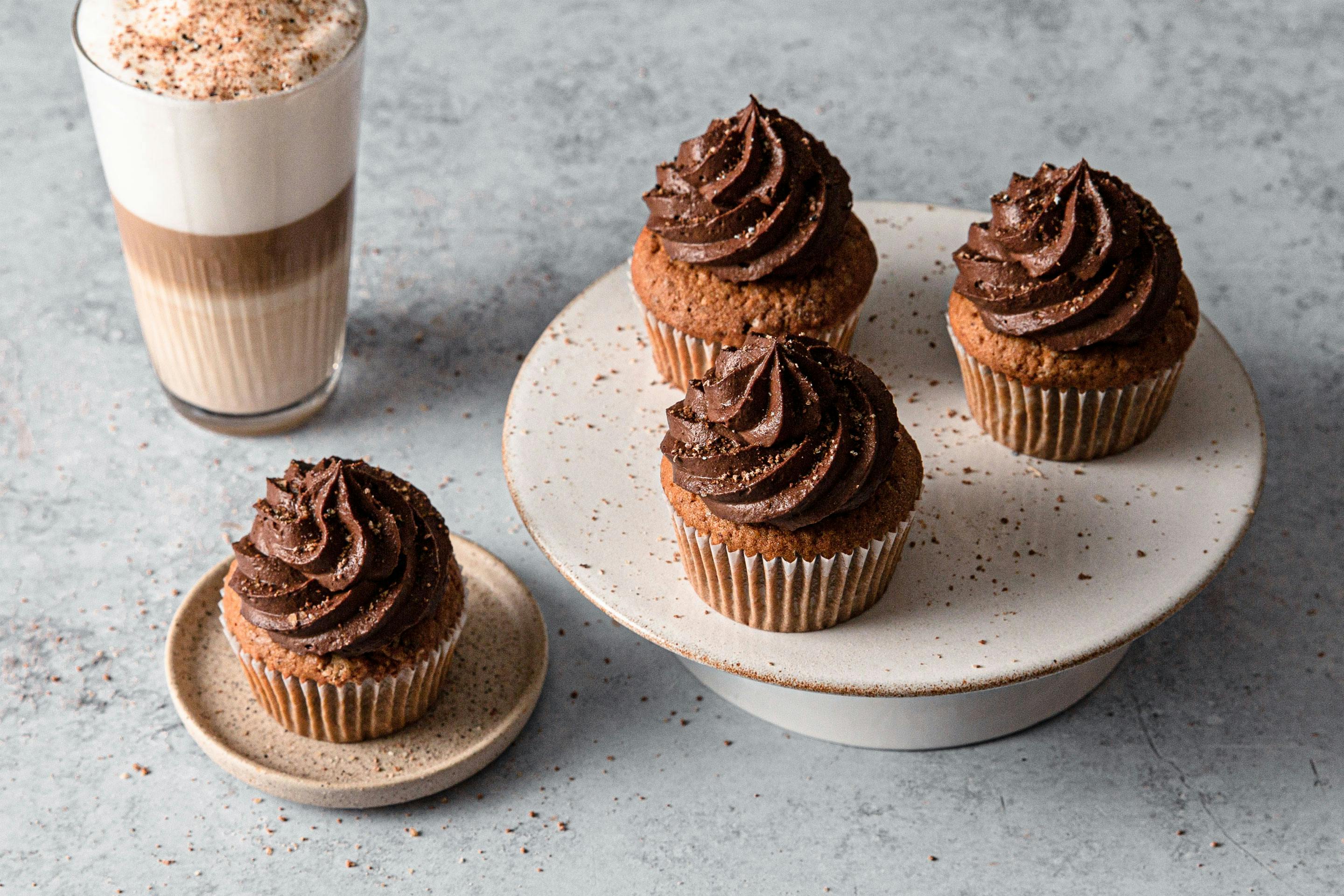 Hazelnut muffins with a dark chocolate topping on a cake plate. Next to it is a layered latte macchiato on a gray work surface.