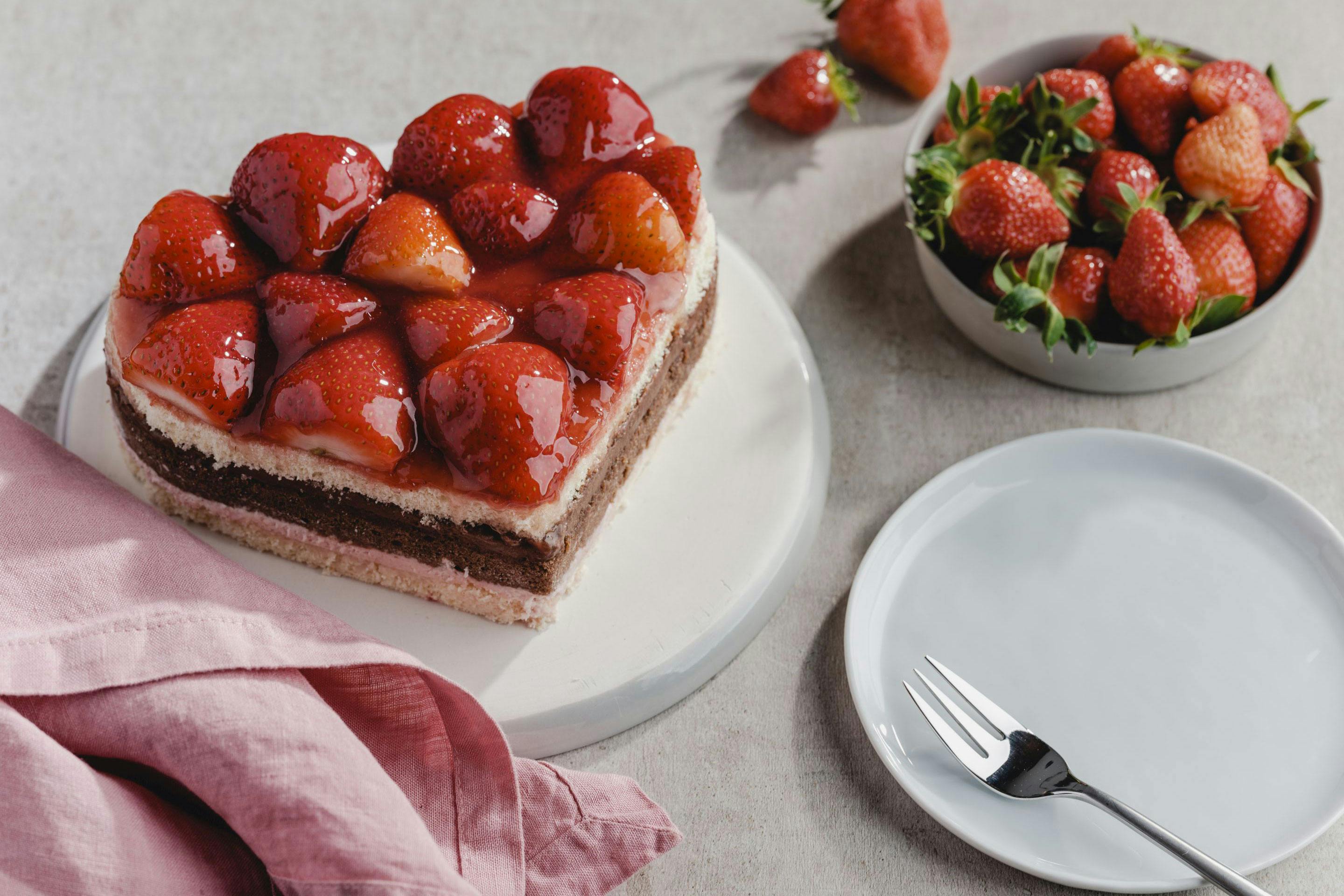 Strawberry chocolate cake in the shape of a heart and a bowl with strawberries as well as a napkin and a plate with a cake fork