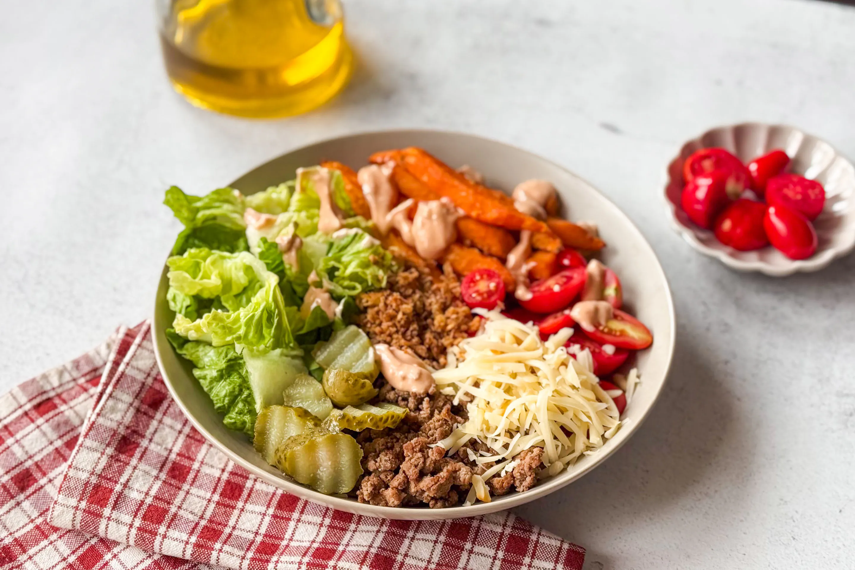 A bowl with romaine lettuce, pickles, ground beef, cheddar cheese, cherry tomatoes, sweet potato fries, and dressing. A small dish with additional cherry tomatoes and a bottle of oil are nearby.