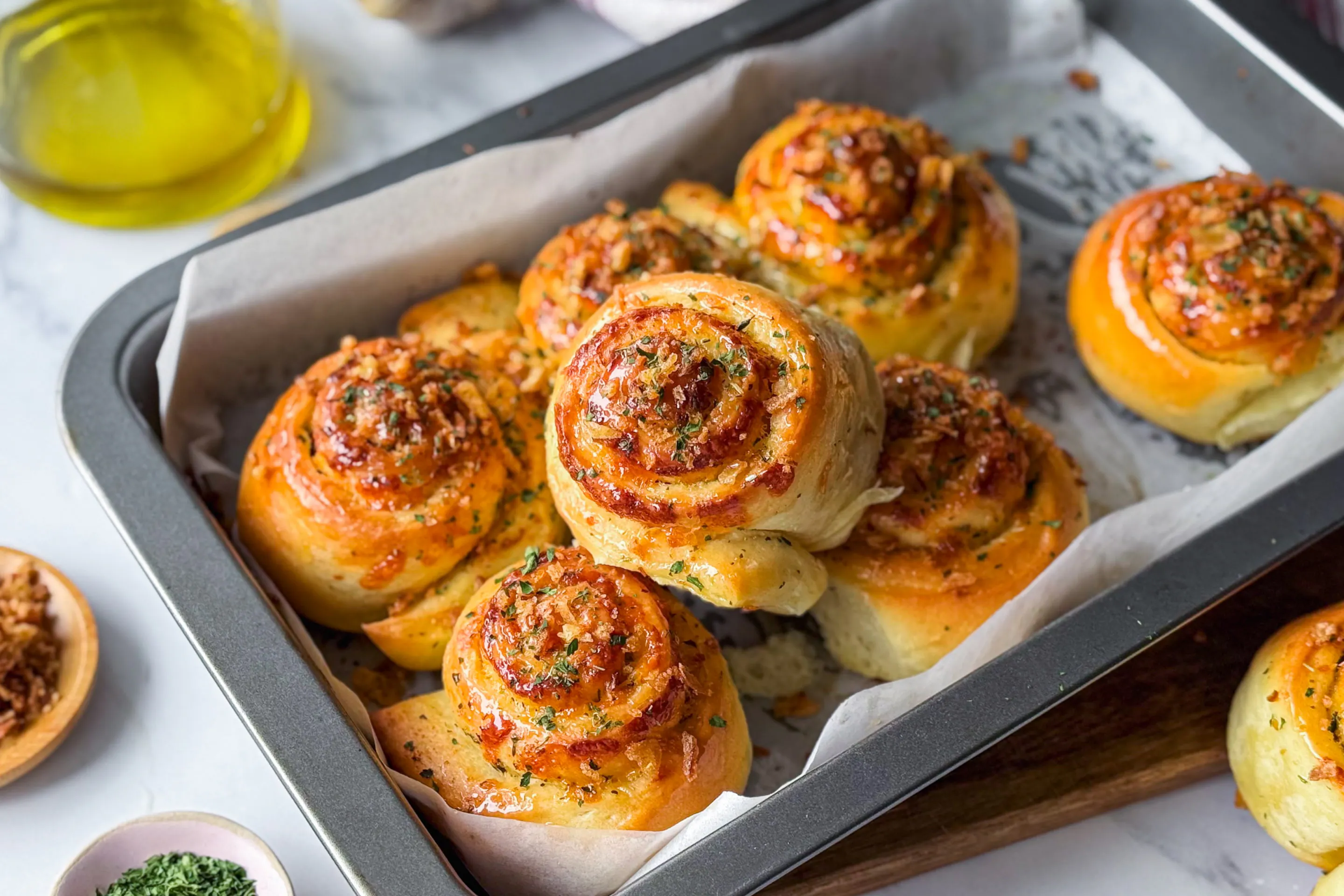 A baking tray filled with freshly baked pizza rolls garnished with herbs and cheese. A bottle of olive oil and small bowls of dried onions and herbs are nearby.