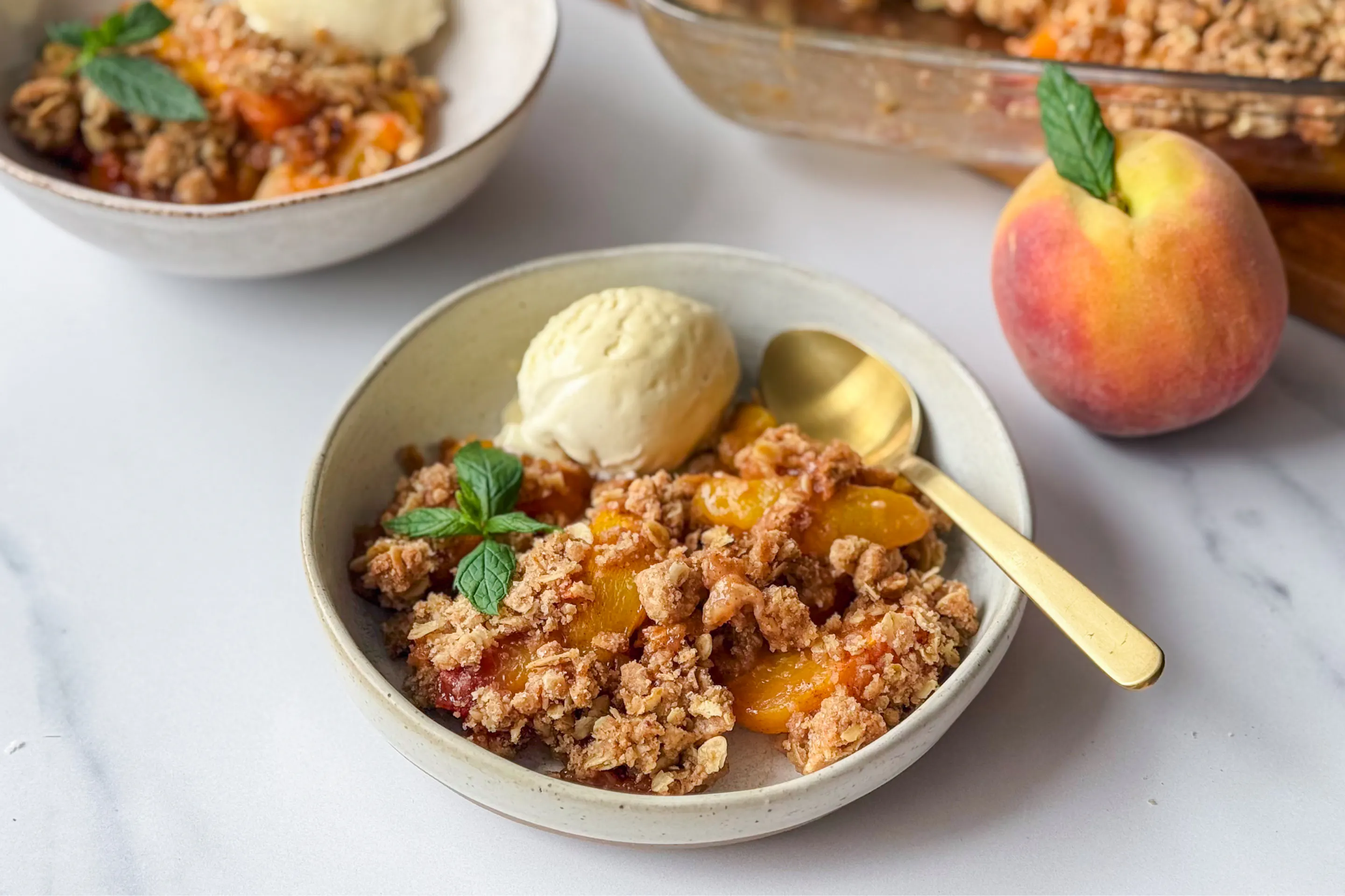 A peach crumble dessert served with vanilla ice cream and garnished with mint leaves, displayed in a baking dish and bowls.