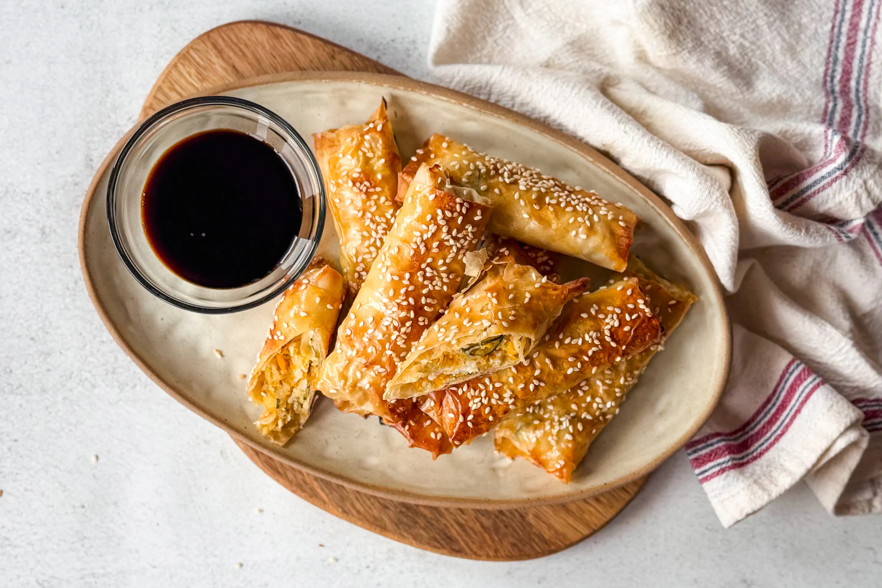 A plate of crispy spring rolls topped with sesame seeds, served with a side of soy sauce. The rolls are filled with a vegetable mixture including carrots and cabbage.